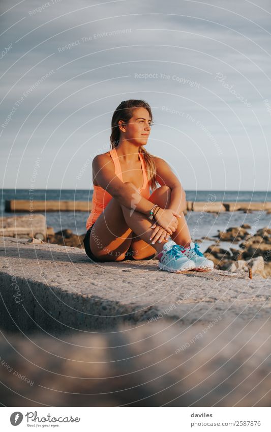 Beautiful woman with sports clothes, sitting on a concrete wall outdoors at sunset. Lifestyle Joy Wellness Relaxation Sun Ocean Sports Fitness Sports Training