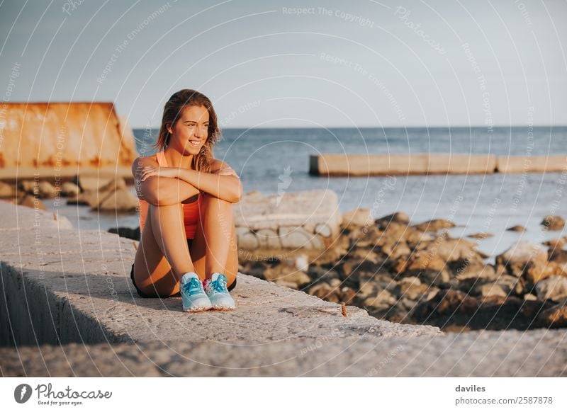 Smiling woman with sports clothes, sitting on a concrete wall outdoors at sunset. Lifestyle Joy Happy Beautiful Body Relaxation Leisure and hobbies Ocean Sports