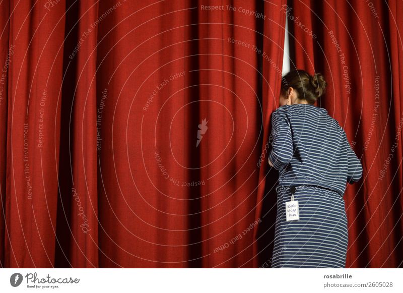 young woman in a blue dress dares to take a look behind the scenes and curiously looks behind a red theatre curtain as a symbol for background information, insights or announcements