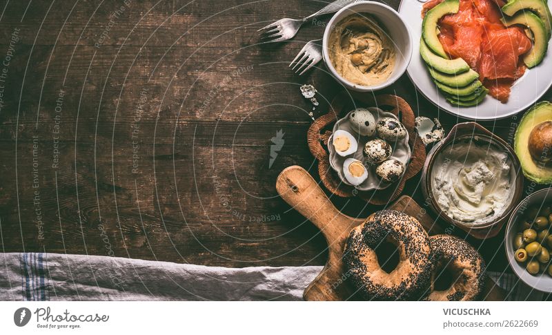 Home breakfast preparation with bagel bread, salmon, avocado , fresh cheese, hummus and cooked quail eggs on dark rustic wooden background, top view. Copy space for your design, banner