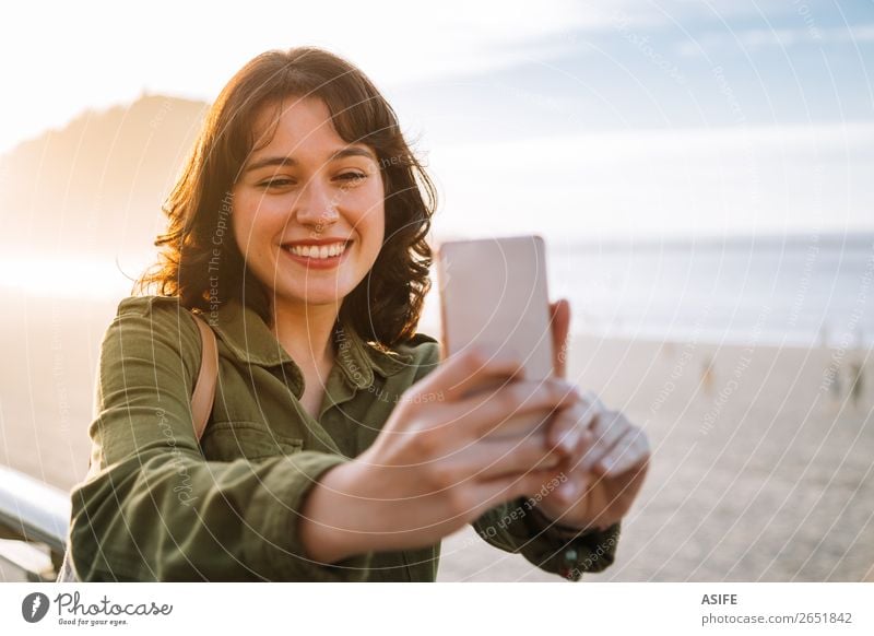 Young happy woman with green jacket taking selfie with her smartphone on the beach at sunset Happy Beautiful Leisure and hobbies Vacation & Travel Beach Ocean