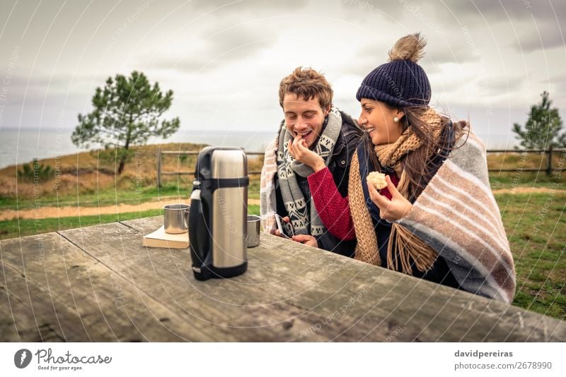 Young couple under blanket eating muffin outdoors in a cold day Eating Beverage Coffee Lifestyle Happy Ocean Winter Table Woman Adults Man Couple Nature Sky