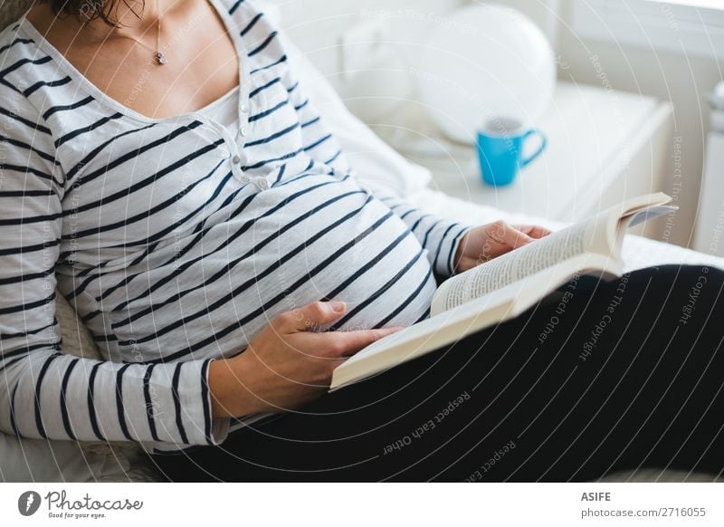 Happy pregnant woman in striped t-shirt sitting on the bed with legs on a cushion reading books about pregnancy, close up Beautiful Body Relaxation