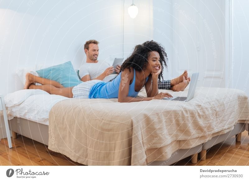 Happy young couple relaxed at home lying down in bed on the tablet and computer interracial Relationship Couple Youth (Young adults) Bed Bedroom Technology