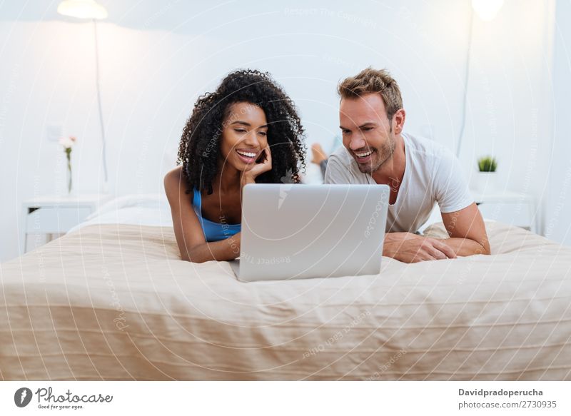Happy young couple relaxed at home lying down in bed on the computer interracial Relationship Couple Youth (Young adults) Bed Bedroom Technology Computer