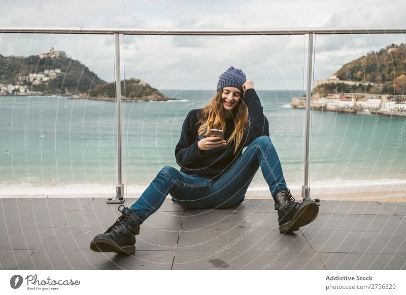 Woman sitting with smartphone at seaside Youth (Young adults) Coast Ocean Provocative Smiling PDA Sit Handrail using pretty Attractive Hat Nature Water