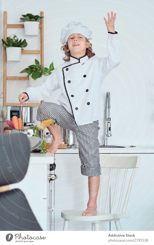 Boy in cook hat with upped hand near pot on chair in kitchen Cook Boy (child) Pot Hand Kitchen Chair chef Child Vegetable Hat Cooking Modern Funny Home Light