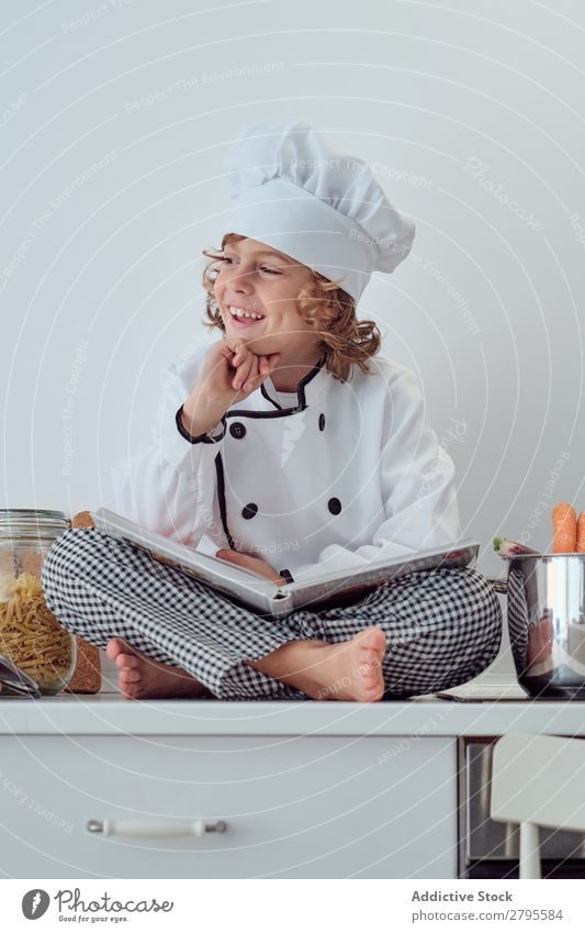 Boy in cook hat with book sitting near pots on electric fryer in kitchen Cook Boy (child) Book Pot Kitchen chef Child Vegetable Hat Volume Reading Stove & Oven