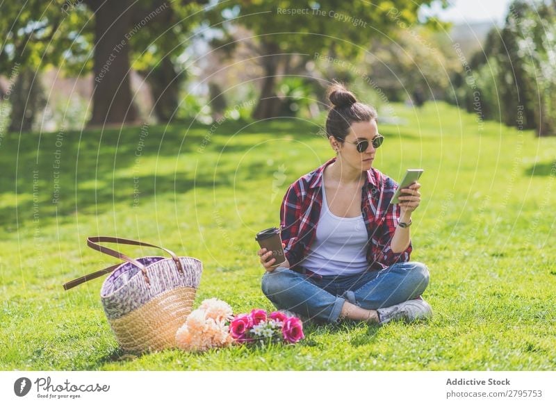 Front view of a young hipster woman wearing sunglasses, sitting on grass in a park while using a mobile phone Young woman Hipster Hip & trendy Sit Park Grass