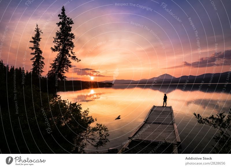 Anonymous traveler on pier in mountains Human being Jetty Sunset Lake Mountain Vacation & Travel Tourist Silhouette Reflection Sky Calm Bright Mirror Forest