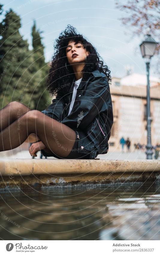 Young woman posing at fountain Woman Attractive City fashionable Curly Brunette Jacket Fountain Water Looking into the camera Fashion Youth (Young adults)