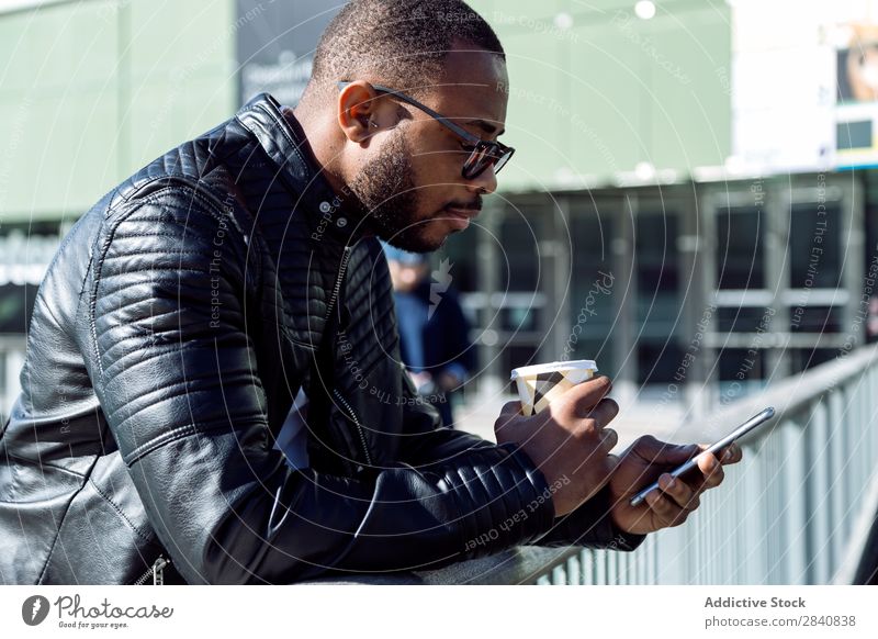 Side view of young man standing with coffee and phone near metal fence. male successful stylish confidence businessman technology morning using browsing manager