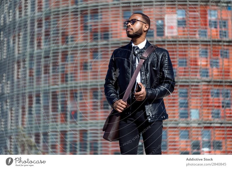 Trendy man in black leather coat and glasses holding smart phone and looking away on urban background. male successful stylish confidence businessman technology