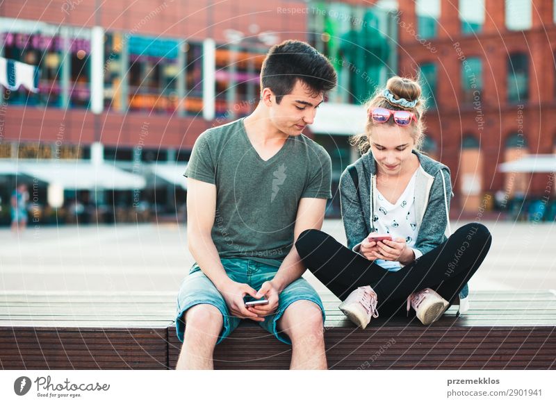 Couple of friends, teenage girl and boy, having fun together with smartphones, sitting in center of town, spending time together Lifestyle Summer To talk