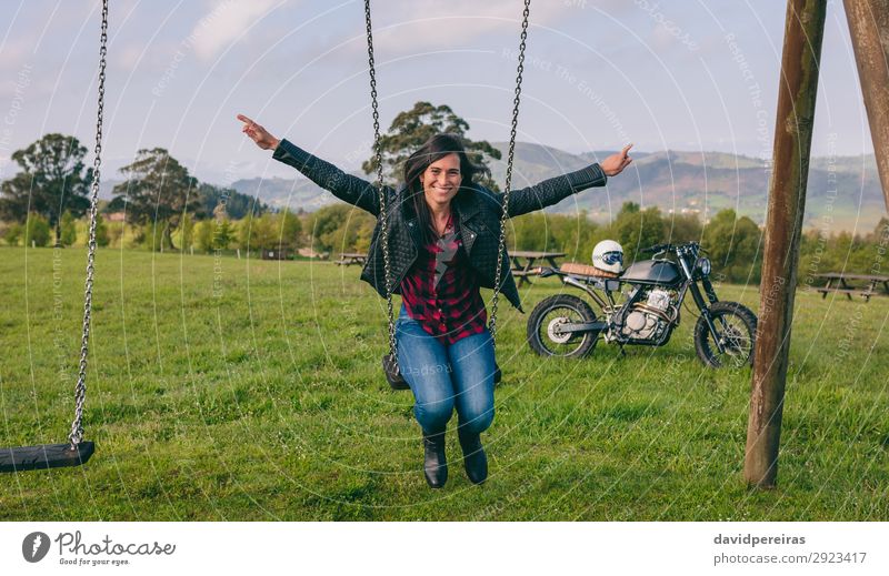 Young woman swinging in recreational area with motorcycle in background Lifestyle Happy Beautiful Freedom Human being Woman Adults Nature Tree Grass Park Meadow