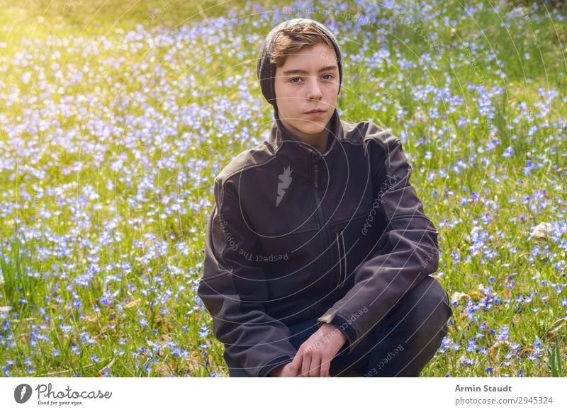 portrait of a boy crouching on a spring meadow with many flowers male young outdoor sunlight blue nature grass green park caucasian cap serious teenager looking