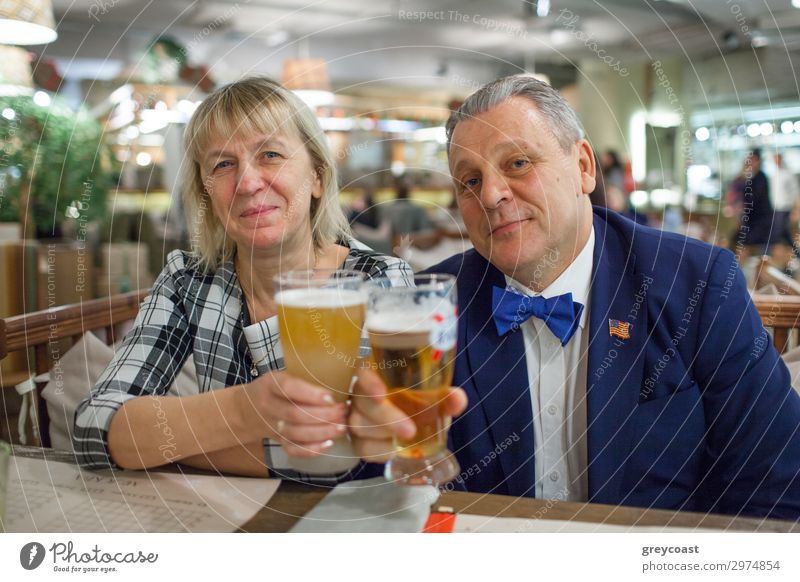A portrait of a middle aged couple sitting at the table close to each other. Man is wearing a blue jacket and a bright blue bow tie. A woman is in a checkered shirt. Both of them are raising glasses of beer and smiling