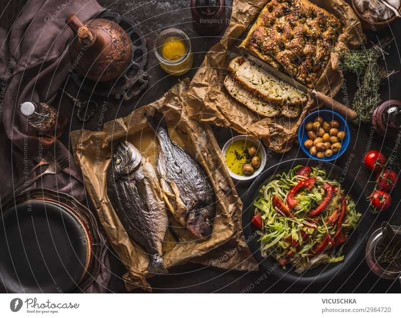 Mediterranean lunch or dinner with roasted dorado fishes, homemade focaccia bread , olive oil and olives served on rustic table with tableware and kitchen utensils, top view.