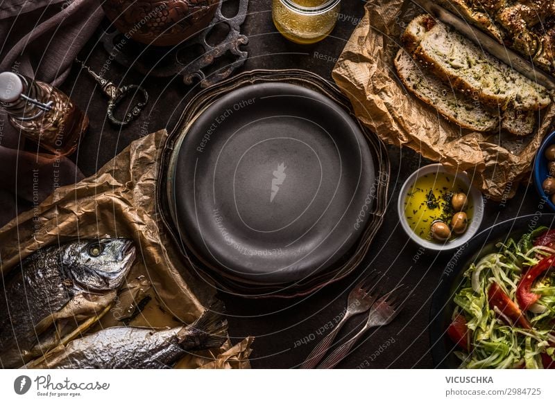 Food background of  Mediterranean food with Focaccia bread , roasted dorado fishes, salad bowl and olives oil on dark rustic kitchen table around empty plate. Delicious Mediterranean  cuisine concept.