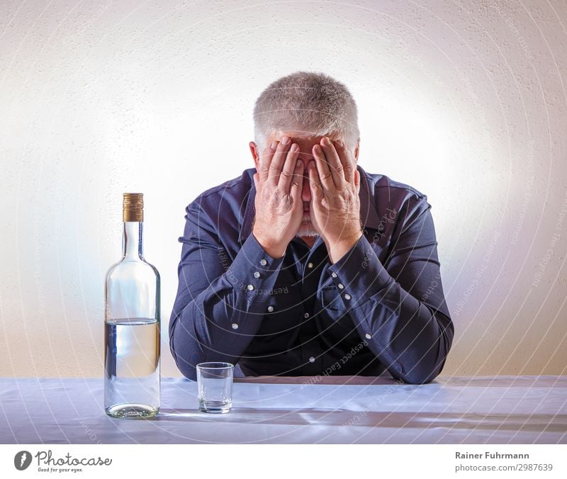 A man sits at a table and covers his face with his hands. On the table is a bottle of alcohol and an empty shot glass Man Alcoholic drinks vodka Alcoholics