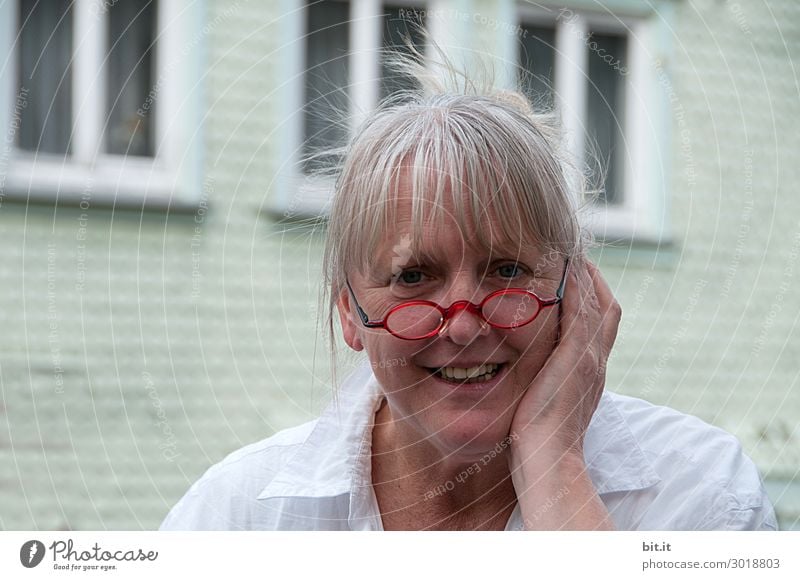 Mature woman with red glasses on her nose, bangs, grey hair, stands in front of a white house with windows. Female senior, with pain in ear or tooth, holds hand protectively against cheek of face. Lady with a pleased, surprised expression.