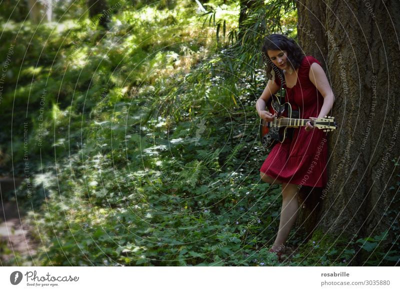 young woman in a red dress plays guitar leaning against a tree in the forest | whom the muse kisses Relaxation Leisure and hobbies Playing Music Young woman