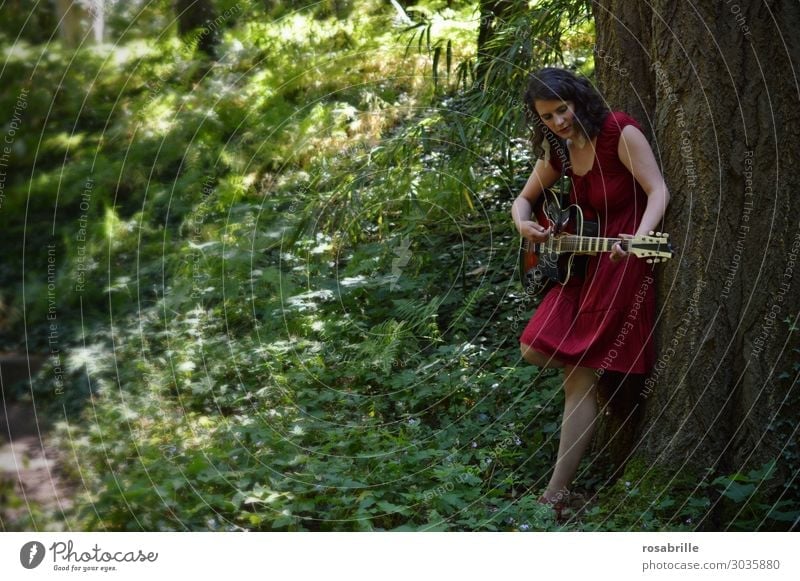 young woman in a red dress plays guitar leaning against a tree in the forest | whom the muse kisses Relaxation Leisure and hobbies Playing Music Young woman