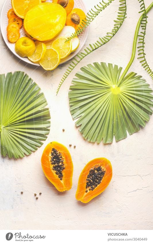 Halved ripe papaya fruit with seeds on white table with tropical leaves and plate with yellow sliced fruits, top view. Summer food. Healthy eating. Breakfast fruits plate. Modern. Copy space