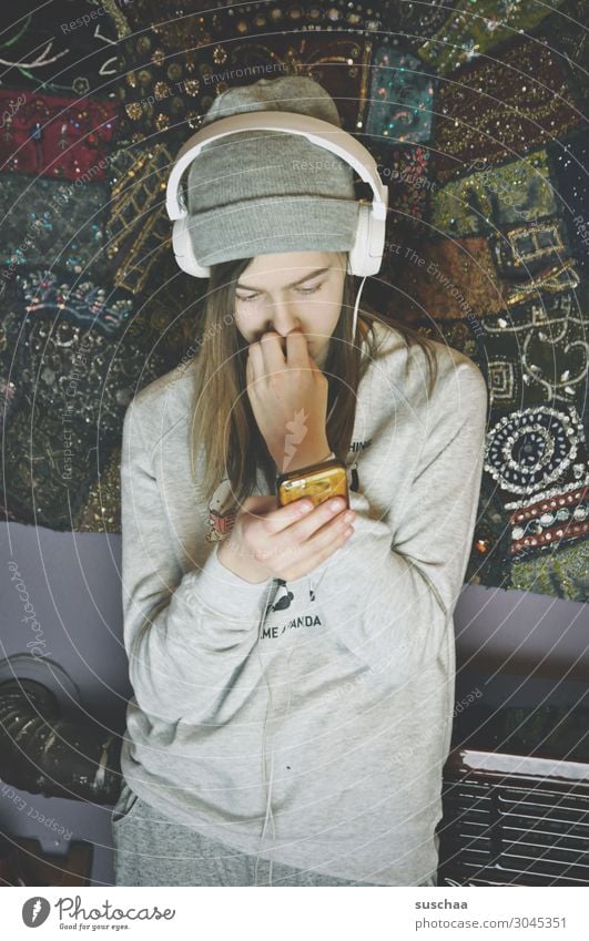 teenager with headphones and cap stands at a wall with an oven in the background and looks at her smartphone Youth (Young adults) girl Living or residing mobile