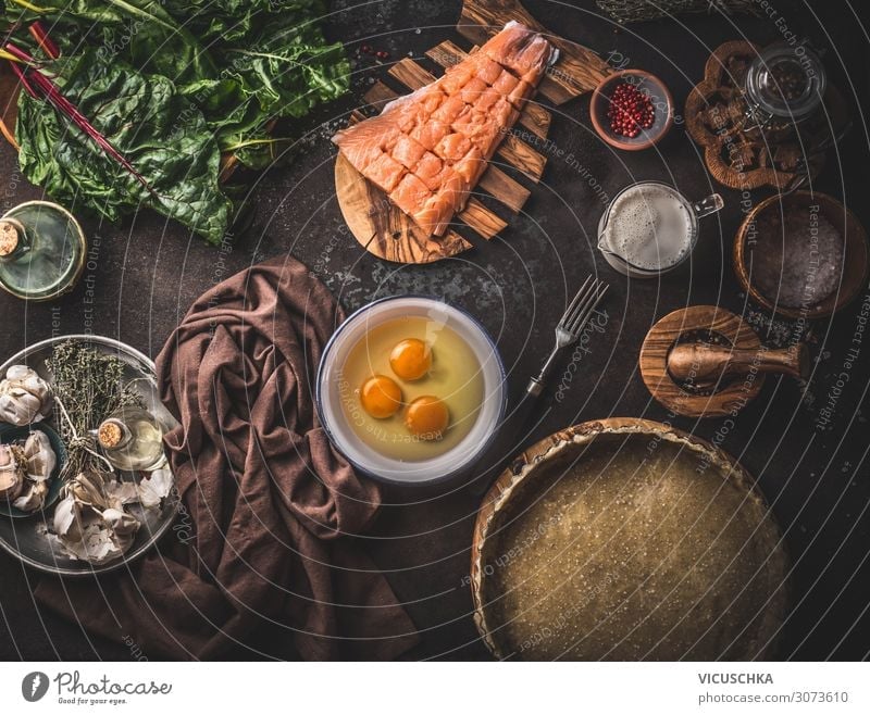 Quiche or open faced pie ingredients: salmon, chard, eggs, cream, dough in baking form on dark rustic table background with kitchen utensils, top view. Cooking preparation