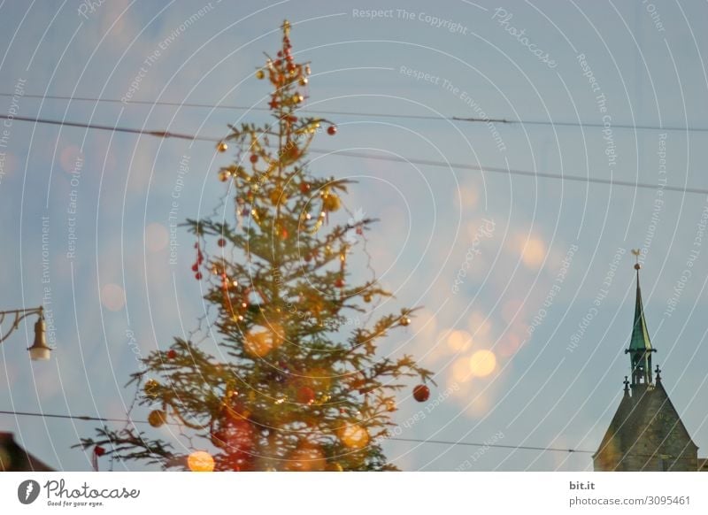 Kitschy, bizarre, decorated Christmas tree stands between wires, church, lamp and lantern in the city and lights up. Funny, abstract, bright, funny, urban Christmas mood with Christmas tree & light outside in front of blue sky in Advent