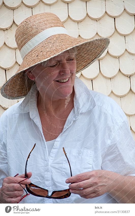 A mature woman with a straw hat on her head, holding a bright white blouse and sunglasses in her hand, is standing in the primordial course in front of an old house with white shingles. Lady is standing in front of a bright wall during a trip through the old town and smiles happily sideways down.