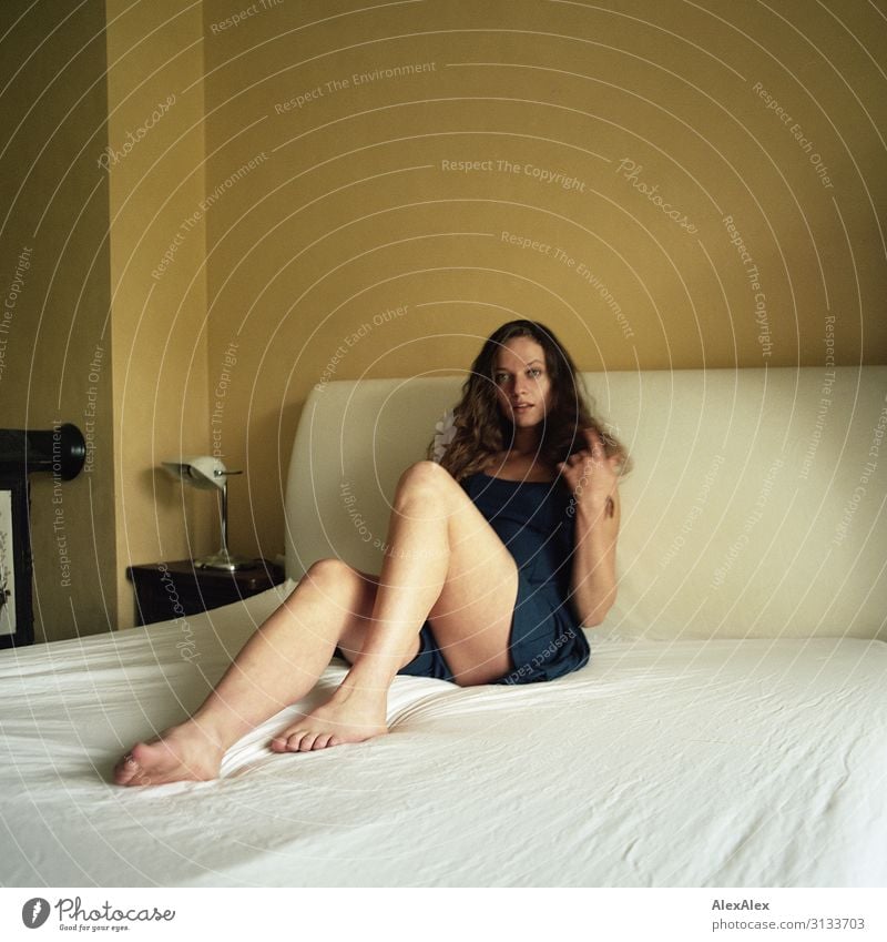 Young woman with long brunette hair and long legs sits on bed in