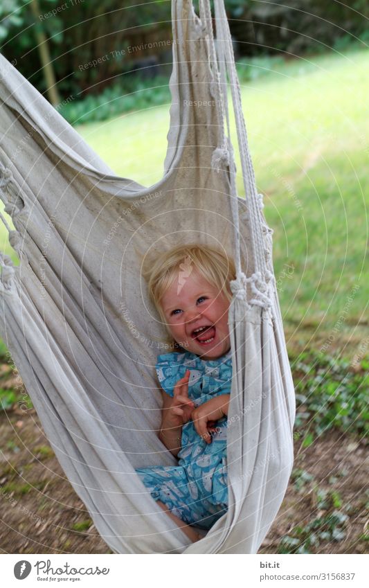 Little, sweet, blonde girl sits, lies happy and beaming in a hammock for children in the garden and laughs heartily into the camera. Leisure and hobbies Playing
