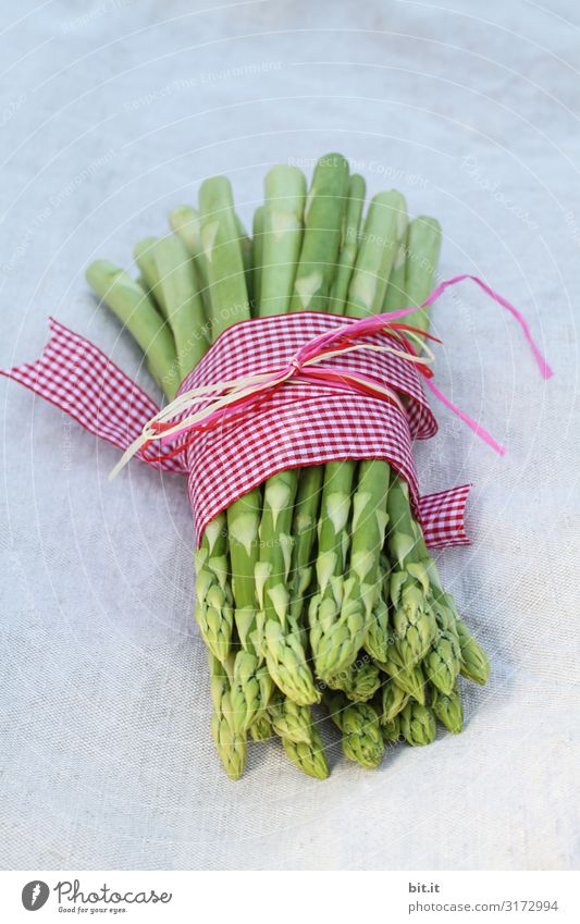 A portion of green asparagus, freshly harvested from the local field, lies on the table, decorated with a checkered ribbon of fabric, tied together, in the light, with a white linen cloth.
