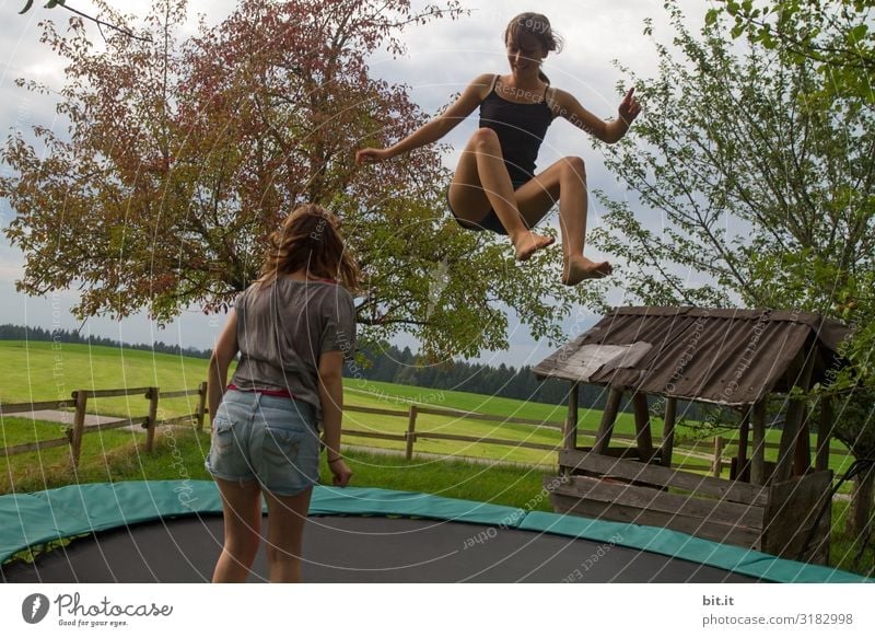 Two girls are jumping on a trampoline together, in nature. One flies up, the sister stands to watch, observe and learn. Sports Fitness Young woman