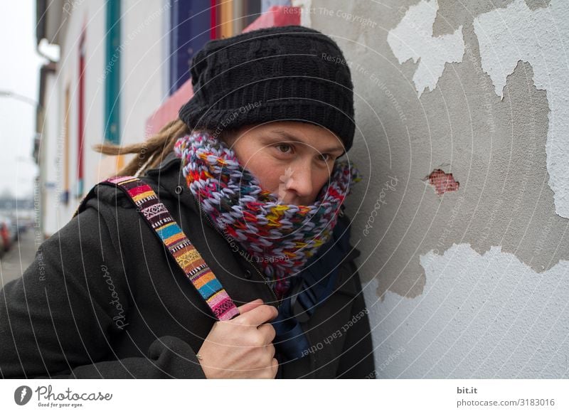 Front view of a young woman with dreadlocks, cap, colourful scarf and bag, who is tired, exhausted, sadly leaning against an old, grey house wall with a colourful window during an excursion in the city, to have a quiet break.