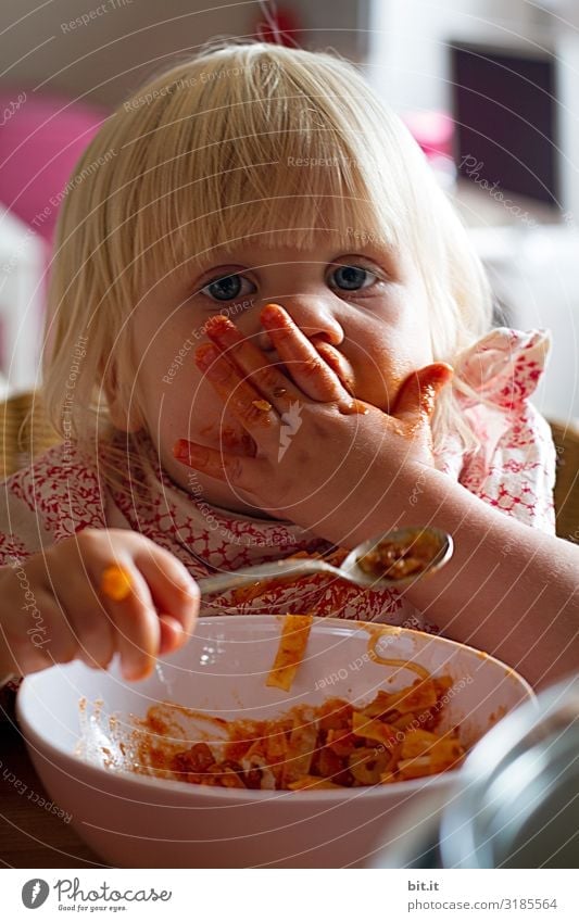 Little blonde sweet girl is noodles with tomato sauce and smears her face with red sauce with her hand, she holds the small spoon in her hand, over a bowl and looks curiously into the camera, at home in the apartment.