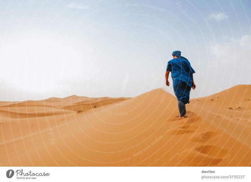 Arabian man in blue clothes walking on a desert dune. - a Royalty