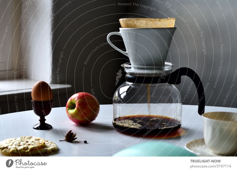 Glass coffeepot, white porcelain filter, light brown filter paper and dark brown continuous bubble forming coffee, apple, egg in wooden egg cup, white cup, corn waffles, white table in front of grey wall in the morning light.