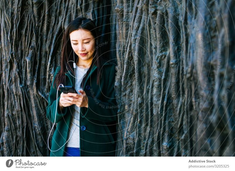 Asian female listening to music near rough wall Woman Listening Music PDA using Wall (building) Lean Root Tree Street City asian Ethnic Rough Relief Surface
