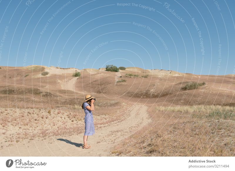 Young female photographer on dry field walk woman road sand landscape camera trail unpaved dusty grass sunny solitude summer straw hat dress casual modern