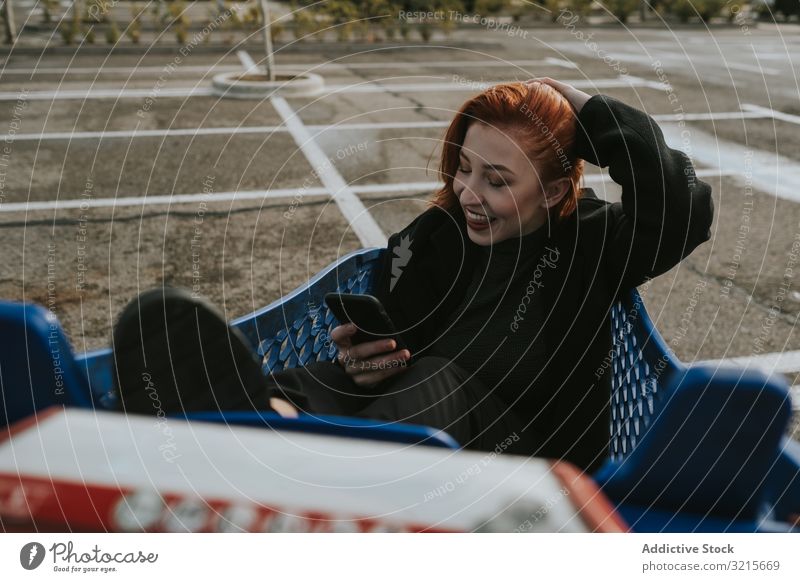 Smiling woman with smartphone in shopping trolley in parking lot selfie attractive taking selfie young beautiful casual cheerful smiling network telephone