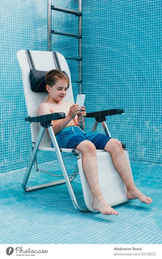 Boy with smartphone sitting on bottom of empty pool boy kid resting chair tile decorated technology gadget device mobile play child vacation childhood game
