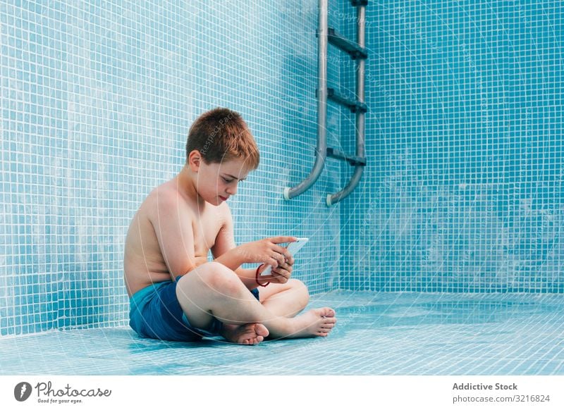 Boy with smartphone sitting on bottom of empty pool boy kid resting tile decorated technology gadget device mobile play child vacation childhood game online