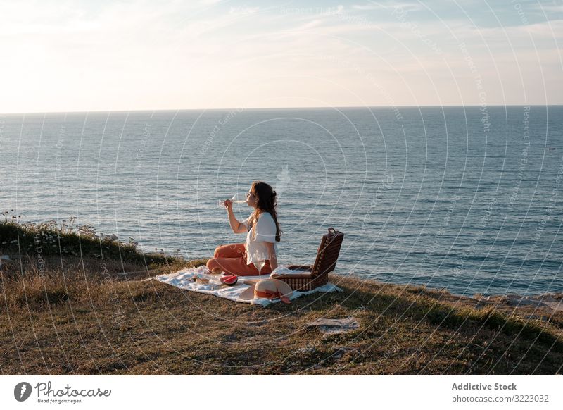 Woman with glass of drink on picnic mat looking at sea and mountains woman seaside beach summer reading leisure sky relax vacation summertime vintage fashion