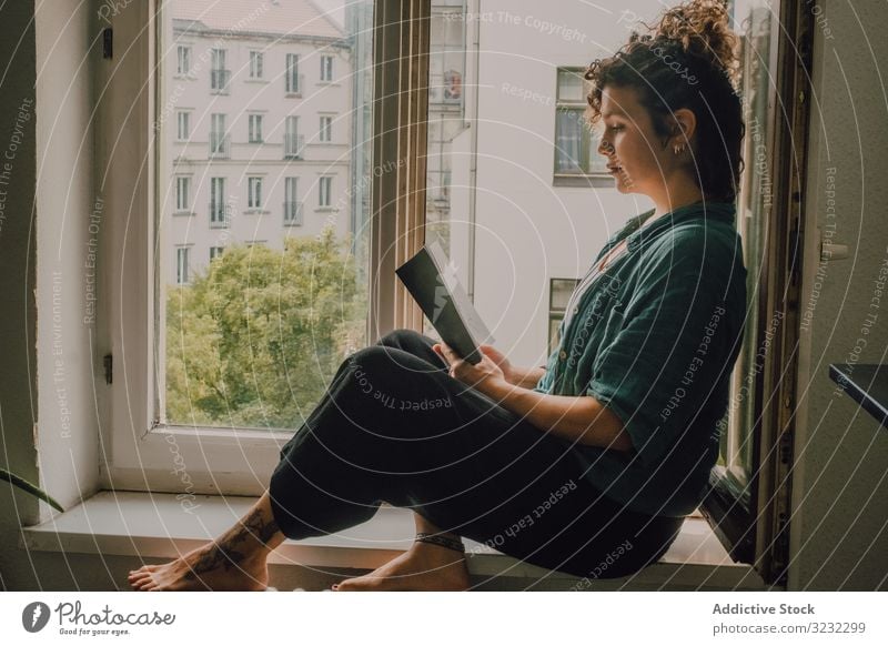 Interested woman reading book at window sill at home interested relaxed literature windowsill barefoot casual sit apartment happy resting young adult peaceful