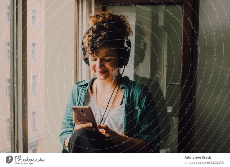 Cheerful woman in headphones using smartphone at home cheerful smile listen happy music browsing window sill curly attractive peaceful relaxed apartment young