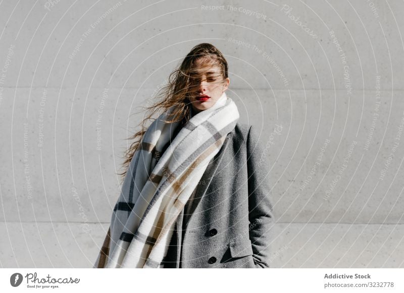 Young woman in coat and scarf on windy day stylish street closed eyes wall city building female urban fashion cool young model outfit warm weather exterior lady