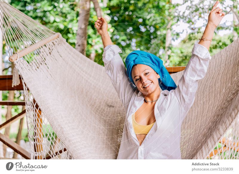 Pleased woman in turban resting in hammock on terrace happy smile raising hands sit tree greenery shirt peaceful costa rica young relax summer comfortable